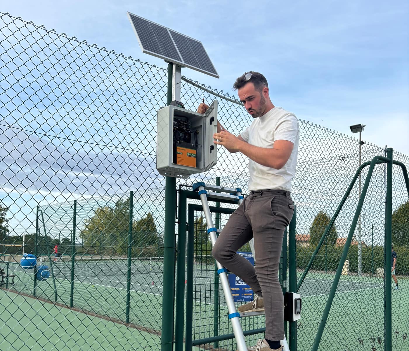 Technicien Neop sur un escabeau installant un boîtier de contrôle d’accès à panneau solaire au portillon d’un court de tennis