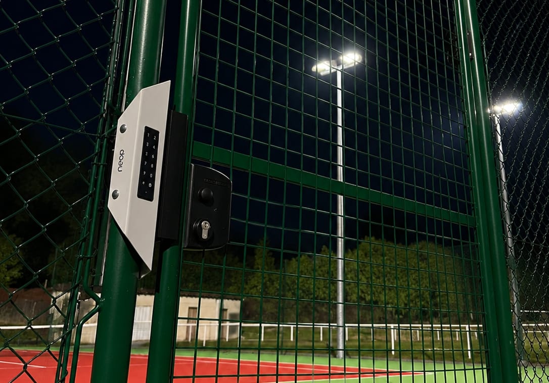 Court de tennis éclairé de nuit par ses projecteurs, avec le clavier d’accès Neop sur le portillon